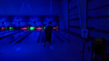 Neon ışıklı kapalı bowling salonu. Bir kişi bovling maçının ortasında, topu kulvardan aşağı atıyor. Atmosfer dinamik ve modern, sağında dur işareti var.
