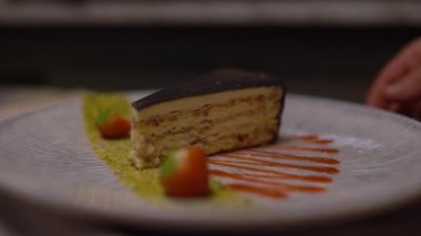 Tabakta bir dilim katmanlı kek, sanatsal sos lekeleri ve yeşil toz ile, bir el yavaşça pastayı çevirir.