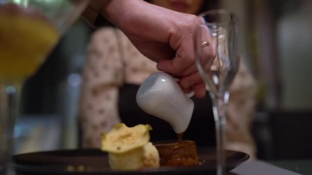皿の上の皿の上にソースを注ぐ手のクローズアップ 明るい背景は 食事の設定を示唆しています ワイングラスは前景にあり 雰囲気は素晴らしいダイニング体験のようだ — ストック動画