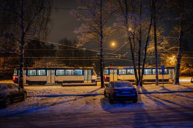 Riga, Letonya - 5 Aralık 2023 - Karla kaplı ağaçlar arasındaki tramvaylı gece sahnesi, sokak ışıklarıyla aydınlatılmış.