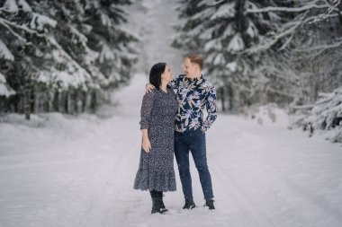 Karda dikilen bir adam ve bir kadın birbirlerine bakıp gülümsüyorlar. Arka planda karlı ağaçlar var..
