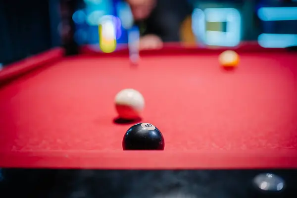 Close-up of billiard balls on red pool table with blurred background, eight ball in focus, player preparing to shoot in the background.