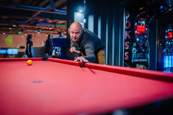 A focused man lines up his shot at pool, eyeing the yellow ball, red table foreground, arcade games brightly lit in the background.