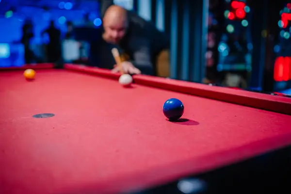 A man focusing intently on a pool shot, cue stick aligned, blue ball in focus, red pool table, blurred background showing arcade ambiance.