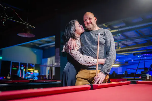woman affectionately embracing a man from behind at a pool table, both smiling and enjoying a moment together in a neon-lit game hall.