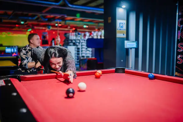 woman focusing on a pool shot with a man in the background, in a vibrant, neon-lit billiards hall.