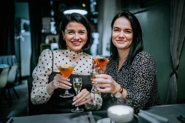 two women in a restaurant smiling and toasting with cocktails, wearing stylish outfits with polka dots and floral patterns.
