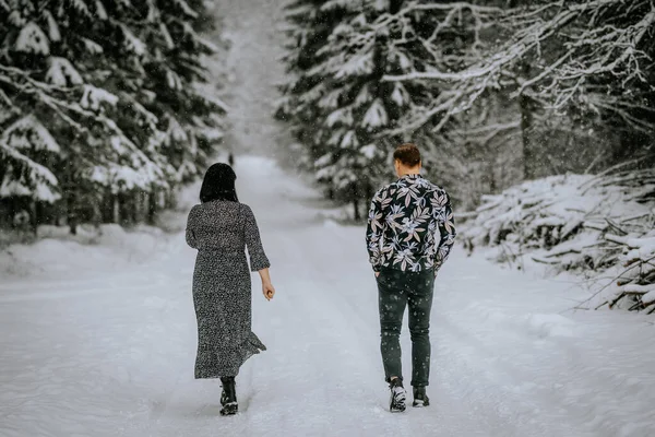 man and a woman walking away on a snow-covered road, surrounded by snowy trees.
