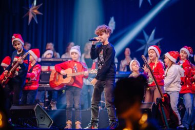 Valmiera, Letonya - 28 Aralık 2023 - Genç, kıvırcık saçlı bir çocukla canlı bir Noel konseri, mikrofona şarkı söyleyen çocuklar gitar çalıyor, bazıları Noel Baba şapkası takıyor.