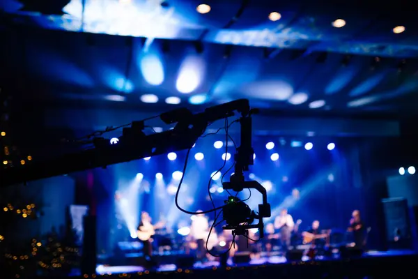 camera crane in the foreground with a blurred band performing on stage under blue stage lighting.