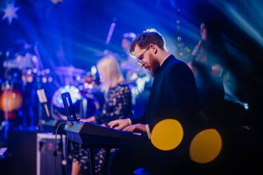 Valmiera, Letonya - 28 Aralık 2023 - Sahnede mavi sahne aydınlatma ve bokeh, arka planda grup üyeleri ile çalan odaklanmış erkek klavyeci.