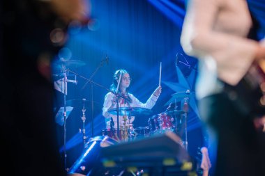 Valmiera, Letonya - 28 Aralık 2023 - Kadın davulcu sahnede davul çalıyor, mavi sahne ışıkları ve diğer müzisyenler yumuşak odaklı arka planda.