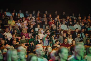 Valmiera, Letonya - 28 Aralık 2023 - Bir etkinliği izleyen kalabalığın arasında kamera operatörü, odaklanmış bir ön planla loş ışıklı bir mekanda görüntülendi ve bulanık bir seyirci kitlesi oluşturuldu..