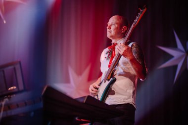 Valmiera, Letonya - 28 Aralık 2023 - Beyaz gömlekli bas gitarist, şenlik kravatı takıyor, sahne arkasında kırmızı bir fon ile şarkı söylüyor..