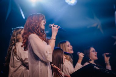 Valmiera, Letonya - 28 Aralık 2023 - Sahnede mikrofona şarkı söyleyen kızıl saçlı kadın, arka planda diğer vokalistlerle birlikte