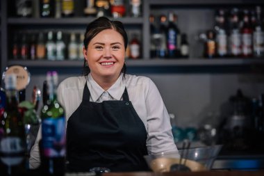 Riga, Letonya - 6 Aralık 2023 - Beyaz gömlek ve siyah önlük giyen gülümseyen barmen, çeşitli şişelerle dolu bir barın önünde duruyor..