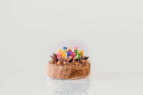 a chocolate cake with happy birthday in colorful candles on a white background with a reflection on the surface