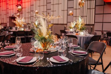 Zarifçe hazırlanmış masa örtüsü, altın çiçekli masa örtüsü ve arka planda daha süslü masalar bulunan masa takımı..
