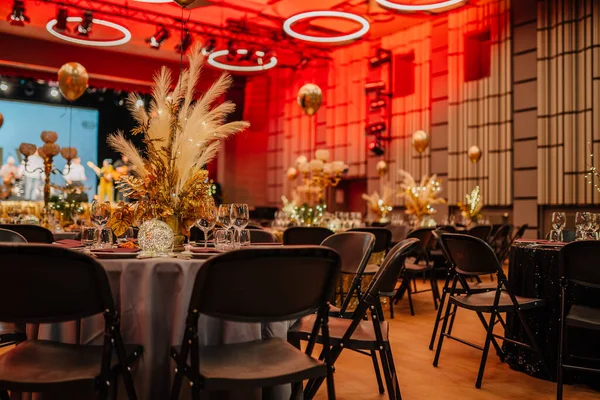a gala event setting with decorated tables, golden centerpieces, and a stage with red lighting and circular decorations in the background.