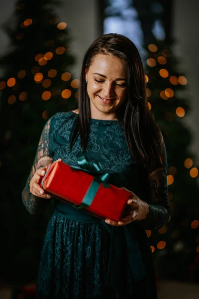 woman in a teal lace dress looks down with a smile at a red gift box with a green ribbon she\'s holding, with a Christmas tree and lights in the background.