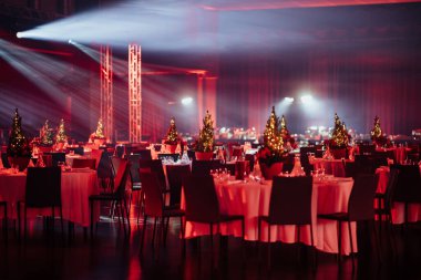 Büyük loş ışıklı etkinlik salonu, akşam yemeği için hazırlanmış birden fazla masa, her biri küçük ışıklandırmalı Noel ağacı süsü ile, bir tavanın altında, tiyatral ışıklandırma ile.