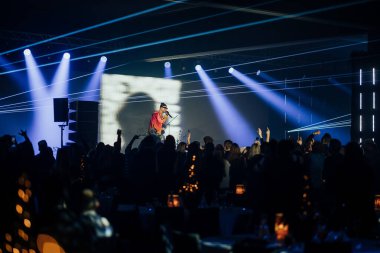 Riga, Letonya - 18 Aralık 2023 - Sahnede mikrofonlu bir sanatçı, seyirciler ellerini kaldırıyor ve arka planda canlı sahne ışıkları var..