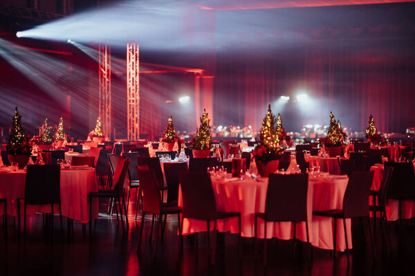 large dimly lit event hall with multiple tables set for a dinner, each with a small lit Christmas tree centerpiece, under a ceiling with theatrical lighting.