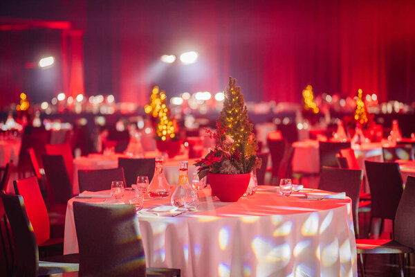 warmly lit event hall with dining tables, Christmas tree centerpieces, and bright spotlights in the background.