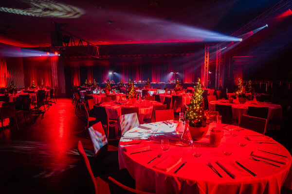 banquet hall set for an event with tables, Christmas tree centerpieces, and dramatic red and blue overhead lighting.