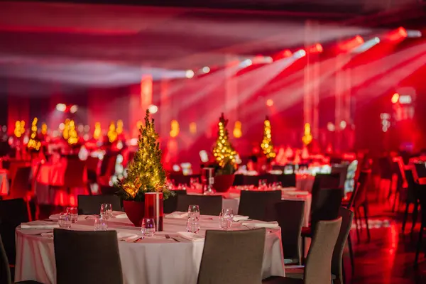 close-up of a banquet table with a small lit Christmas tree centerpiece in a hall with ambient red lighting and more tables in the background.