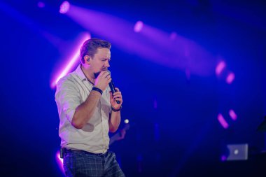 Riga, Letonya - 18 Aralık 2023 - Erkek, arka planda mavi ve mor ışıklandırmayla sahnede yoğun duygularla bir mikrofona şarkı söylüyor.