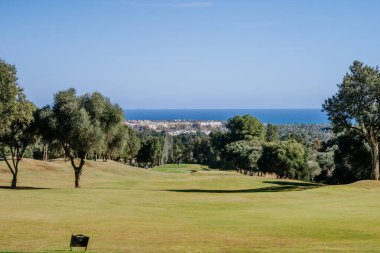 Malaga, İspanya - 21 Ocak 2024 - Deniz manzaralı bir golf sahası Ağaçlar, açık bir gökyüzü ve uzak bir şehir manzarası.
