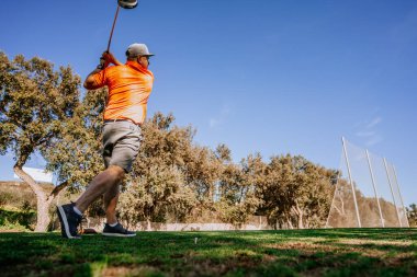 Malaga, İspanya - 21 Ocak 2024 Turuncu gömlekli bir golfçü golf sahasındaki açık gökyüzü ve ağaçlardan oluşan bir zemine doğru sallanırken yakalandı.