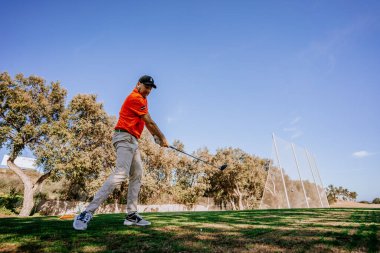 Malaga, İspanya - 21 Ocak 2024 Turuncu gömlekli bir golfçü golf sahasındaki açık gökyüzü ve ağaçlardan oluşan bir zemine doğru sallanırken yakalandı.