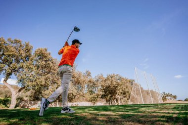 Malaga, İspanya - 21 Ocak 2024 Turuncu gömlekli bir golfçü golf sahasındaki açık gökyüzü ve ağaçlardan oluşan bir zemine doğru sallanırken yakalandı.