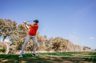 Malaga, İspanya - 21 Ocak 2024 Turuncu gömlekli bir golfçü golf sahasındaki açık gökyüzü ve ağaçlardan oluşan bir zemine doğru sallanırken yakalandı.