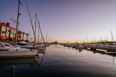 Sotogrande, İspanya - 21 Ocak 2024 - Limanda demirli yelkenliler, sakin sular, palmiye ağaçları ve suya yansıyan renkli bir gökyüzü.