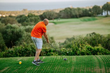 Malaga, İspanya - 21 Ocak 2024 Turuncu gömlekli ve beyaz şortlu kel bir adam deniz manzaralı bir golf sahasında salıncakta sallanıyor..