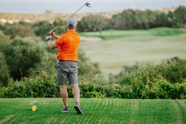 Malaga, İspanya - 21 Ocak 2024 - Turuncu tişörtlü ve gri şortlu bir adam yemyeşil bir sahada manzaralı bir arka planda golf sopası sallamaya hazırlanıyor.