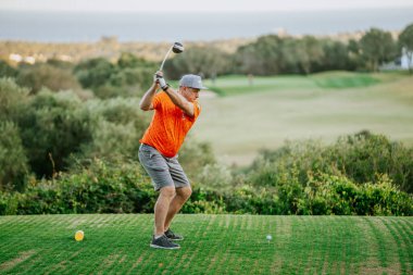 Malaga, İspanya - 21 Ocak 2024 - Turuncu tişörtlü ve gri şortlu bir adam yemyeşil bir sahada manzaralı bir arka planda golf sopası sallamaya hazırlanıyor.