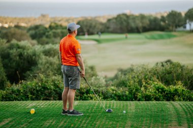 Malaga, İspanya - 21 Ocak 2024 - Turuncu tişörtlü ve gri şortlu bir adam yemyeşil bir sahada manzaralı bir arka planda golf sopası sallamaya hazırlanıyor.