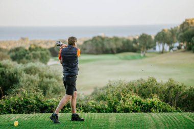 Malaga, İspanya - 21 Ocak 2024 - Siyah şortlu ve turuncu ve siyah yelekli bir adam denize bakan bir rotada golf topuna vurmak üzere.