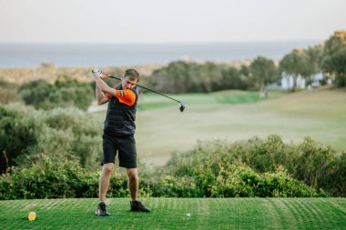 Malaga, İspanya - 21 Ocak 2024 - Siyah şortlu ve turuncu ve siyah yelekli bir adam denize bakan bir rotada golf topuna vurmak üzere.