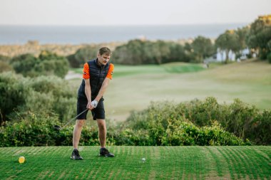 Malaga, İspanya - 21 Ocak 2024 - Siyah şortlu ve turuncu ve siyah yelekli bir adam denize bakan bir rotada golf topuna vurmak üzere.