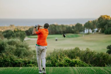 Malaga, İspanya - 21 Ocak 2024 Turuncu tişörtlü bir golfçü, deniz manzaralı ve açık gökyüzü olan bir golf sahasında sallanıyor..