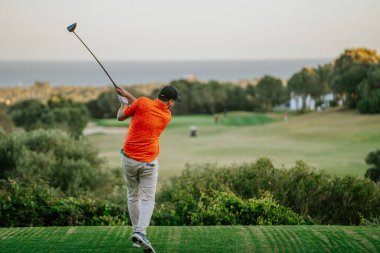 Malaga, İspanya - 21 Ocak 2024 Turuncu tişörtlü bir golfçü, deniz manzaralı ve açık gökyüzü olan bir golf sahasında sallanıyor..