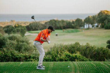Malaga, İspanya - 21 Ocak 2024 Turuncu tişörtlü bir golfçü, deniz manzaralı ve açık gökyüzü olan bir golf sahasında sallanıyor..