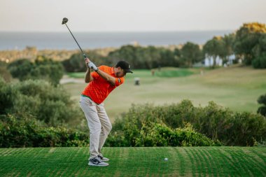 Malaga, İspanya - 21 Ocak 2024 Turuncu tişörtlü bir golfçü, deniz manzaralı ve açık gökyüzü olan bir golf sahasında sallanıyor..