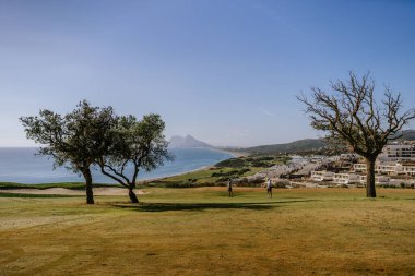 Sotogrande, İspanya - 23 Ocak 2024 - Açık gökyüzü, iki ağaç, golfçüler ve arka planda Cebelitarık Kayası olan kıyı golf sahası.