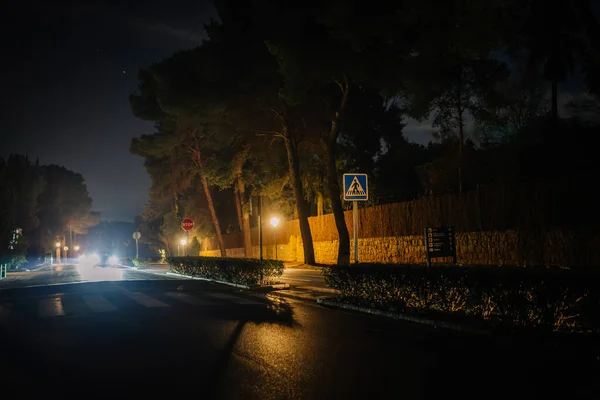 Sotogrande, Spain - January, 23, 2024 - This is a night scene of a street with a pedestrian crossing sign, stop sign, illuminated street lamps, trees, and an approaching car\'s headlights.